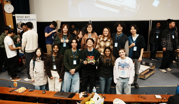 Group of students posing together in a lecture theatre with robotics equipment and competition arena visible in the background.